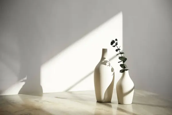 Ilık güneş ışığı, çimen yapraklarından gölgeler, masa üstünde vazo ve fincanlar gibi çeşitli nesnelerin olduğu bir oda.