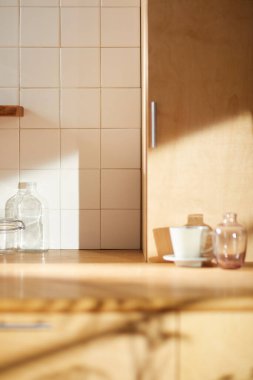 Beyaz fayans duvarlı güneşli bir mutfak, ahşap bir masa ve lavabo..