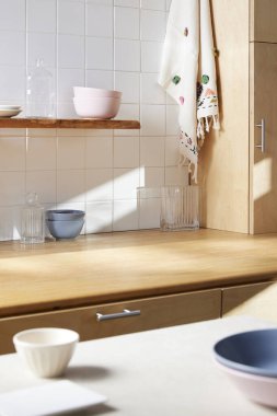 Beyaz fayans duvarlı güneşli bir mutfak, ahşap bir masa ve lavabo..