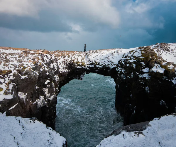 Man walking on a snowy stone arch over the ocean in Arnarstapi, Iceland.