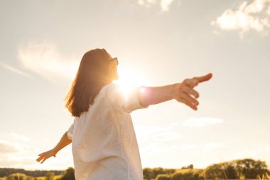 Genç kadın gün batımında çayırda kollarını açmış neşeli ve özgür hissediyor.