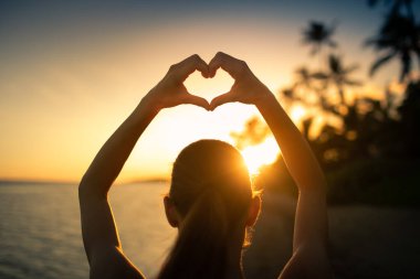 Gün batımında plajda kalp şekillendiren bir kadın.
