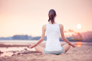 Şafak vakti kumlu bir sahilde, sakin bir sabah ışığı ve sakin bir atmosferle meditasyon yapan bir kadının sakin görüntüsü bir farkındalık ve rahatlama hissi yaratıyor.