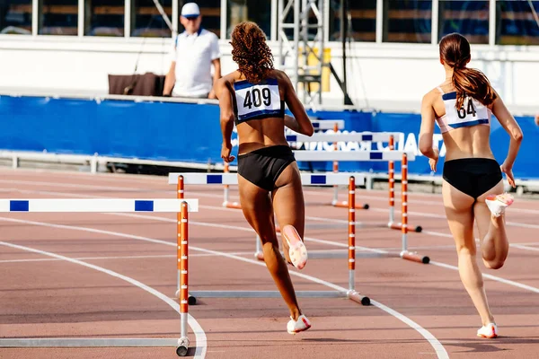 两名女运动员在体育场跑道上跑跨栏 夏季田径锦标赛 黑人和白人女短跑运动员 — 图库照片