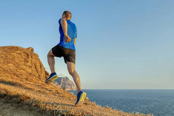 男のランナーが青い空を背景に上り坂を走り夏は海岸沿いを日没でジョギング — ストック写真