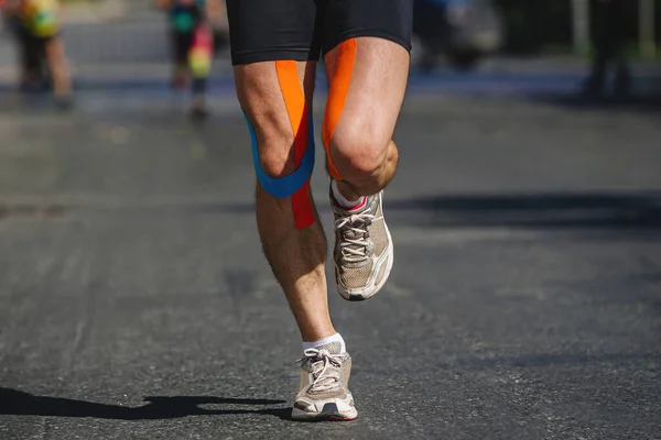 close-up legs male runner running dark asphalt in summer marathon race, kinesiotape on hips and knees