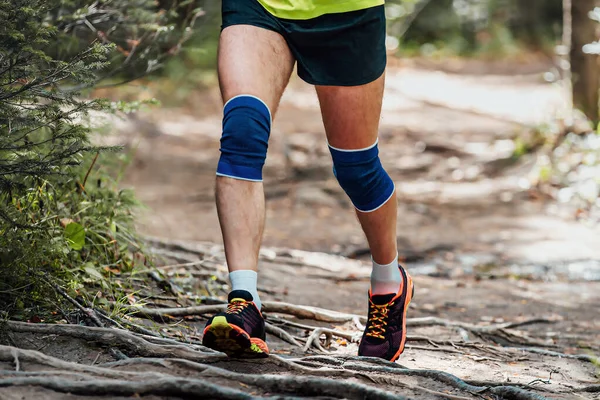 male runner in knee pads running forest trail on tree roots, summer marathon race