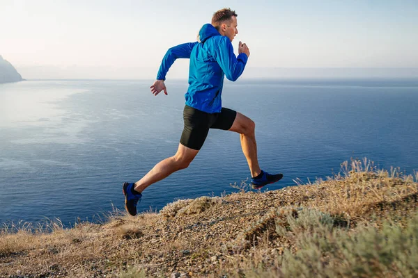 男子跑步者穿着蓝色夹克和黑色紧身衣 背景是天空和大海 沿着山边跑上山路 — 图库照片