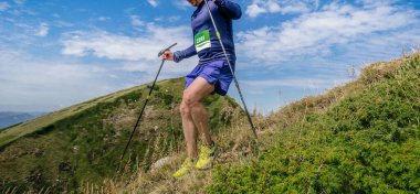 Elinde sırıkla gezen bir sporcu dağ maratonunda dik yamaçtan iniyor.