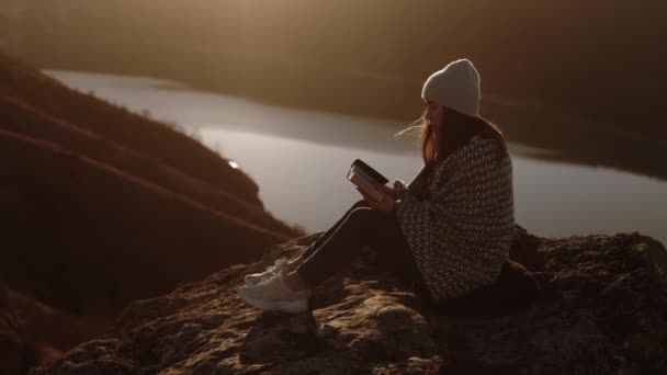 年轻美丽的女人坐在岩石上 夕阳西下 美丽的景色映衬着圣经 欣赏着风景和大自然 — 图库视频影像