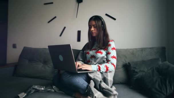年轻迷人的女人穿着休闲装 裹着毛毯 手里拿着笔记本电脑 坐在家里沙发上 头戴耳机 望着窗外 — 图库视频影像
