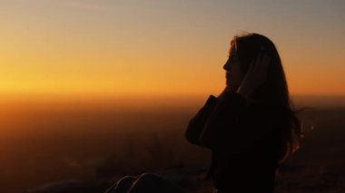 Gün batımında, sarı gökyüzüne karşı gülümseyen mutlu bir kadın silueti. İnanılmaz bir manzarayla dağın tepesinde oturuyor, kulaklık takıyor ve müzik dinliyor, gezgin, açık havada yürüyor.