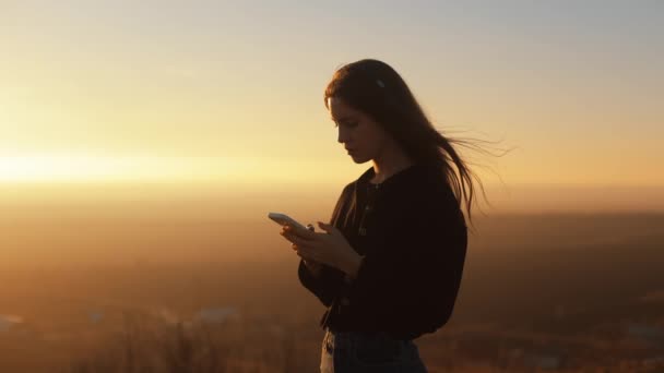 髪の長い若い女性は 黄色の空と美しい風景の背景に夕日に屋外に立っている間 スマートフォンを使用して 風が彼女の髪を吹いて — ストック動画