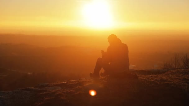 男の観光客のシルエット 彼の手にスマートフォンで日没時に信じられないほどの景色を持つ山の岩の上に座っている男 — ストック動画