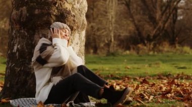 Genç, çekici, kulaklıkla müzik dinleyen bir kız sonbahar parkında düşen yaprakların arasında bir battaniyenin üzerinde oturmuş anın ve müziğin tadını çıkarıyor. Yüksek kalite 4k görüntü