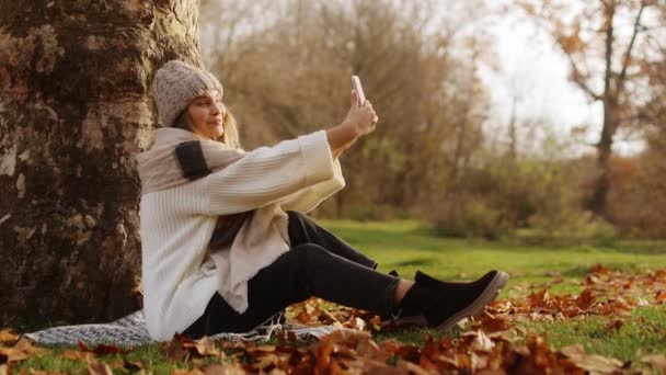 在阳光明媚的一天 小女孩在智能手机的前置摄像头上给自己拍照 在秋天的公园里 坐在一片落叶中的毛毯上 高质量的4K镜头 — 图库视频影像