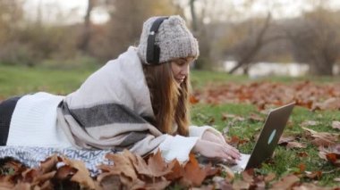 Charming blonde woman has remote work or online study lying on blanket among fallen leaves in yellow autumn park. Smiling young woman with headphones listening to music and working on a laptop. High quality 4k footage