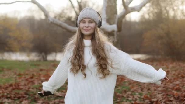 公園で一人で森の中を歩いている帽子のきれいな若い長髪の女性の女性とヘッドフォンで音楽を聞いて 幸せな踊りの旋回 周りを見て 人生を楽しんでいます 高品質4K映像 — ストック動画