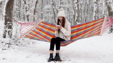 Young woman girl uses a smartphone while sitting in a colorful bright hammock in the middle of a snowy winter park, looks up at the camera and smiles, unusual winter vacation, Christmas vacation. High quality 4k footage