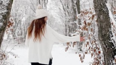 Back view, the camera follows a young beautiful blonde woman in a hat walking through a snowy winter forest, touching frozen tree branches from which snow is falling. silence frosty Christmas wood, sweet face. High quality 4k footage