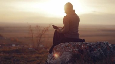  Young woman sitting alone on a rock in a field, reading the bible, praying in nature with an incredible view, watching a beautiful sunset in an autumn evening, peace and tranquility, faith, God's . High quality 4k footage