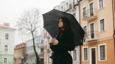 Portrait of a young cute smiling attractive woman girl standing alone on a city street in the rain, holding a black umbrella and drinking coffee through a straw, loves the rain. High quality 4k footage