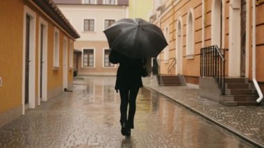 Rear view of young cute smiling attractive stylish woman girl walking alone on city street in rain, holding black umbrella and cup of coffee, turning to camera and smiling. High quality 4k footage