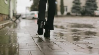 Back view of female slim legs in boots and stylish pants walking on cobblestones on city street in the rain, walking in the rain