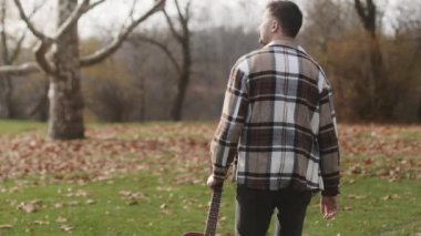 Man with acoustic instrument enjoying nature and sunlight. People, lifestyle, student, expression and style concept. A man in a shirt with a guitar walks on the lawn of an autumn park. High quality 4k footage