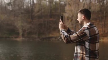 Genç bir adam güzel bir sonbahar fotoğrafı çekiyor göl manzaralı ve telefonuyla park manzaralı. Parkta tek başına yürümek, sonbahar doğasının güzelliğine hayran olmak. Yüksek kalite 4k görüntü