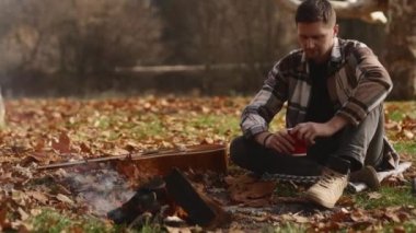 Genç erkek gezgin gülümseyerek ve ateşin yanında kahve içerek sonbahar ormanının çimenliklerindeki bir gitarla ateşe odun atıyor. Açık hava eğlencesi. Yüksek kalite 4k görüntü