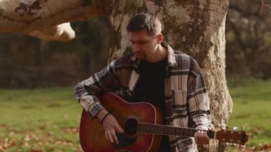 Genç bir adam tek başına gitar çalıyor, dışarıda bir ormanda bir ağacın gövdesine yaslanıyor. Sonbahar manzarası ve sıcaklığı. Yüksek kalite 4k görüntü