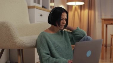 Attractive middle-aged woman in wireless headphones enjoying music, sitting on carpet on floor at home and holding laptop on lap during free time at home, dancing and singing along while working. High quality 4k footage