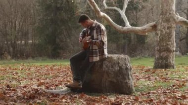 Young man playing guitar sitting on stone in autumn park alone, yellow leaves falling from trees, side view, slow motion