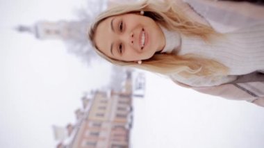 Happy beautiful young woman girl holding smartphone and talking to camera, congratulating on Christmas, filming vlog, making video call standing on snowy street during snowfall. Vertical video.