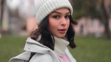 Close-up portrait of charming smiling young woman in warm hat and scarf looking at camera outdoors on background of city street. 