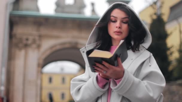 一个女人站在教堂附近看书 一个女孩在户外读圣经 一个女人手里拿着一本圣经 在街上学习上帝的话语 寻找真相 — 图库视频影像