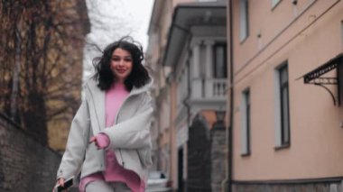 Attractive cheerful woman young brunette girl walking on city street with smartphone in hand, looking at camera and smiling, jumping and twirling, front view