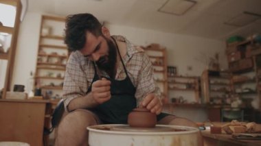 A male potter works on a potter's wheel, making a ceramic pot with a lid from clay in a pottery workshop, a young bearded potter examines his work, corrects imperfections. High quality 4k footage
