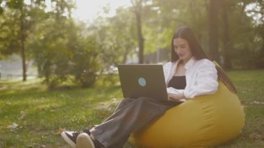 Young woman sit in park on beanbag communicating by video call. Girl speaking looking at laptop computer, online conference distance office chat, virtual training concept IT specialist working outdoor