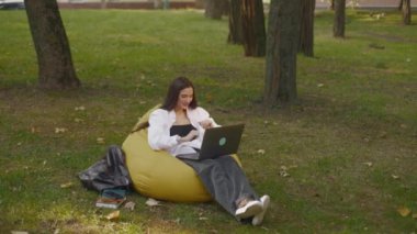 Beautiful brunette girl student in white shirt and jeans talking on laptop by video chat while sitting on beanbag chair in the park. Woman watching webinar on laptop. Education concept. Advertisement