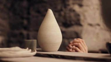Close up. A handmade clay vase sits on a shelf in a pottery workshop. Women's hands take it
