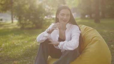 Portrait of a smiling young long-haired brunette woman. Cute stylish girl student sitting in beanbag chair in park in garden near university campus in sunlight. High quality 4k footage