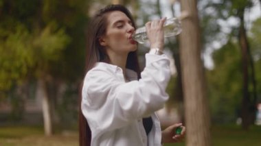 A young woman drinking water from a bottle while walking in the park taking a break to drink water. Young woman long haired brunette drinking water outdoors from bottle. High quality 4k footage