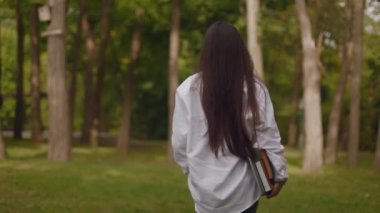 Portraits of a beautiful young woman student girl in a white shirt long-haired brunette in sunglasses holding books textbooks and walking in the park near the campus circle. High quality 4k footage