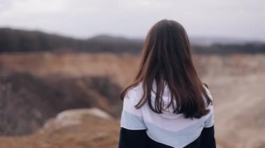 A smiling young woman approaches the top of the mountain peak to the edge of the cliff, raises her hands in the air, happily enjoys the incredible view