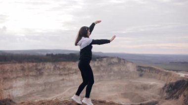 young woman long-haired girl in headphones listening to music and singing along dancing and jumping on the edge of a cliff above a canyon, having fun alone, cheerful tourist