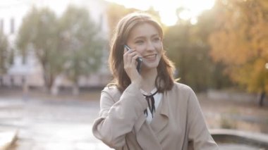 Caucasian confident business woman walking down the street in the city and talking on the mobile phone on the way to the office outdoors. Stylish elegant young woman uses a smartphone. High quality 4k footage