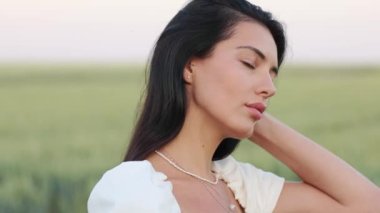 Face of young woman looking at camera, serious look, attractive beautiful hair, brunette, tanned, standing outdoors in a field of wheat, holding a spikelet in her hands, summer girl outdoors natural