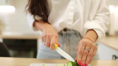 Kotlu genç bir kadın mutfakta sağlıklı bir salata hazırlıyor. Kadın eli kesme tahtasındaki bıçakla salatalığı kesiyor. Bir kadın bir parça salatalık tadıyor. Çevre dostu vejetaryen